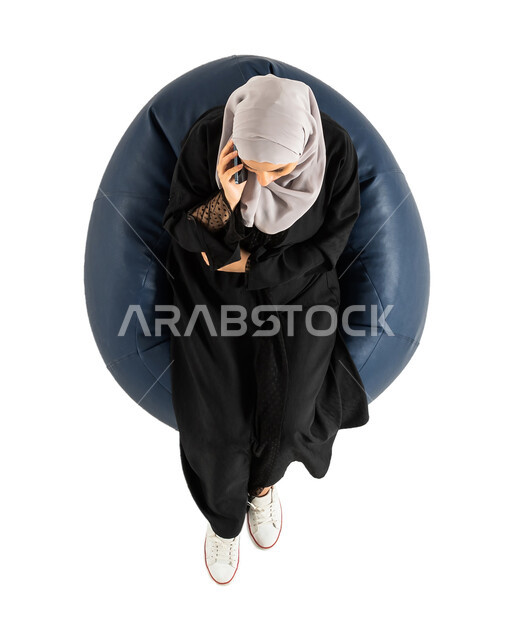 عرض خدمات وتطبيقات الهاتف المحمول، شاشة سوداء فارغة، بورتريه لصورة علوية عامودية لشابة عربية خليجية سعودية تجلس على كرسي وتمسك بيدها الهاتف المحمول ، الجلوس على كرسي مريح ،  التسوق الإلكتروني، خلفية بيضاء