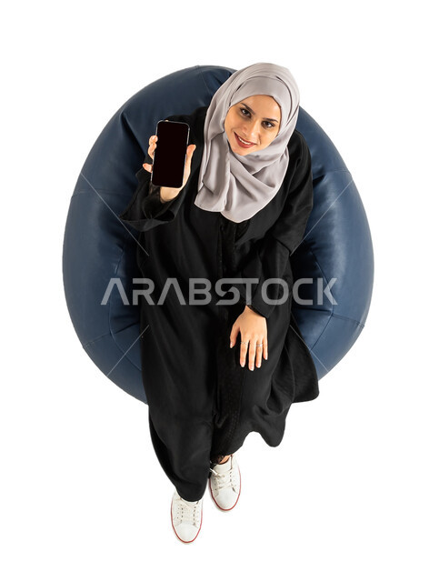 شاشة سوداء فارغة، بورتريه لصورة علوية عامودية لشابة عربية خليجية سعودية تجلس على كرسي وتمسك بيدها الهاتف المحمول ، الجلوس على كرسي مريح ، عرض خدمات وتطبيقات الهاتف المحمول، التسوق الإلكتروني، خلفية بيضاء