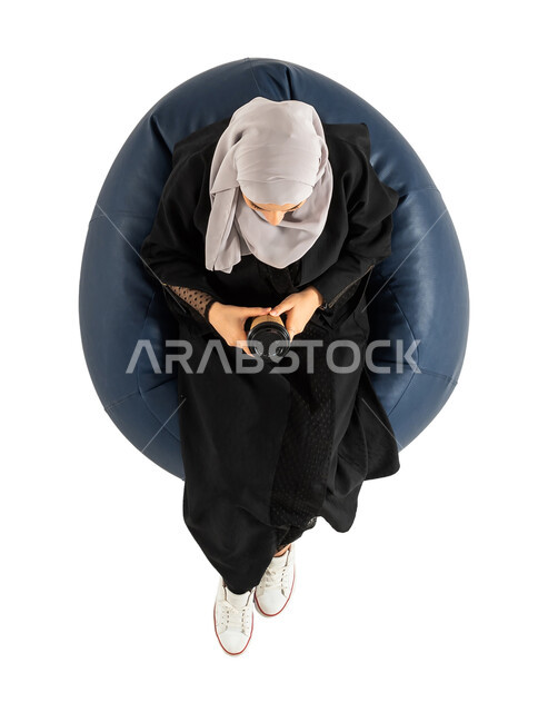 امرأة تمسك بيدها كوب القهوة ، الجلوس على كرسي مريح ، تناول القهوة والمشروبات الساخنة ، بورتريه لصورة علوية عامودية لأمرأة عربية خليجية سعودية تجلس على كرسي ،  خلفية بيضاء