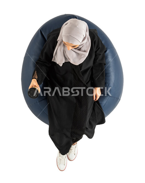 الجلوس على كرسي مريح ، امرأة تمسك بيدها كوب القهوة ، تناول القهوة والمشروبات الساخنة ، بورتريه لصورة علوية عامودية لأمرأة عربية خليجية سعودية تجلس على كرسي ،  خلفية بيضاء