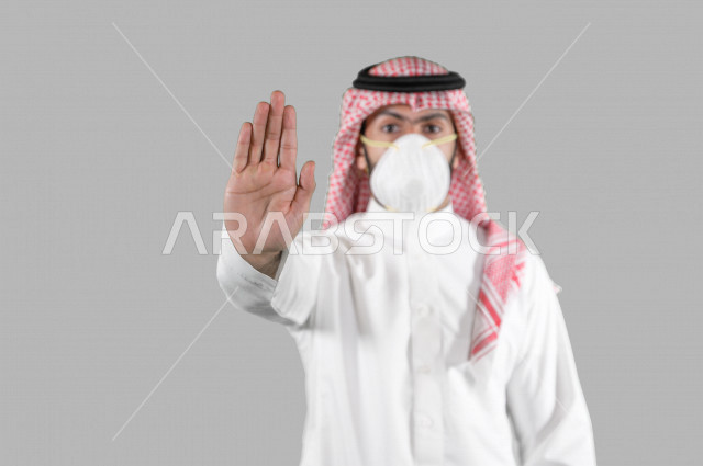 بورتريه لشاب عربي خليجي سعودي يرتدي الكمامة، اتخاذ اجراءات الوقاية من فايروس كورونا (كوفيد-19)، شاب يرفع يده ليتجنب اقتراب الاخرين منه، تجنب انتشار العدوى، خلفية بيضاء