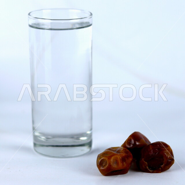 صورة مقربة لحبات من التمر مع كأس من الماء، وجبة ذات قيمة غذائية عالية، ضيافة رمضانية، إفطار رمضان، خلفية بيضاء