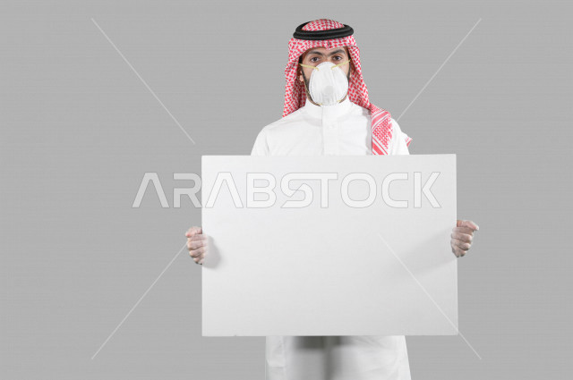 بورتريه لشاب عربي خليجي سعودي يرتدي الكمامة والقفازات، اتخاذ اجراءات الوقاية من فايروس كورونا (كوفيد-19)، شاب يحمل بيده لوحة بيضاء كبيرة، لوحة فنية فارغة، لوح اعلاني أبيض، خلفية بيضاء