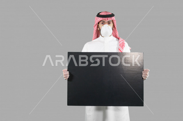 بورتريه لشاب عربي خليجي سعودي يرتدي الكمامة والقفازات، اتخاذ اجراءات الوقاية من فايروس كورونا (كوفيد-19)، شاب يحمل بيده لوحة بيضاء كبيرة، لوحة فنية فارغة، لوح اعلاني أسود، خلفية بيضاء