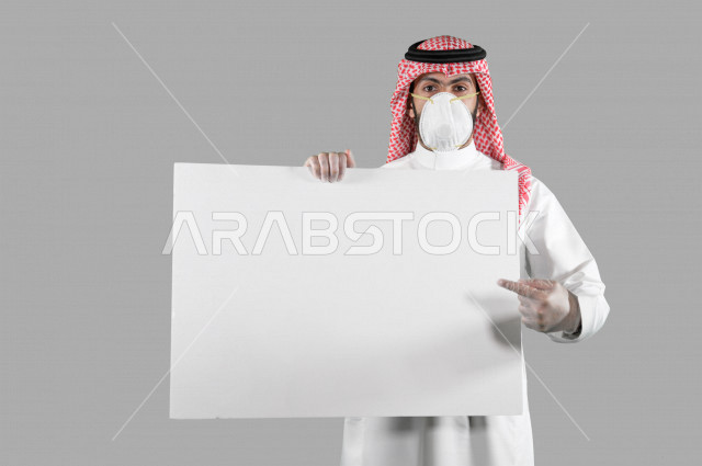 بورتريه لشاب عربي خليجي سعودي يرتدي الكمامة والقفازات، اتخاذ اجراءات الوقاية من فايروس كورونا (كوفيد-19)، شاب يحمل بيده لوحة بيضاء كبيرة، لوحة فنية فارغة، لوح اعلاني أبيض، خلفية بيضاء