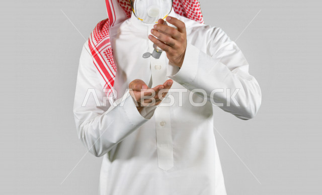 Portrait of a Saudi Arabian Gulf youth, wearing a protective mask, taking measures to prevent corona virus (Covid-19), using hand sanitizer, sterilizing hands, avoiding the spread of infection, white background