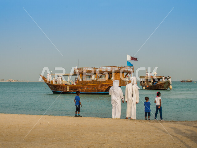 صورة من الخلف لرجلان عربيان خليجيان قطريان يقفان على شاطئ البحر، اطفال قطريين، القوارب الخشبية، علم قطر، مراكب سياحية، السياحة في قطر
