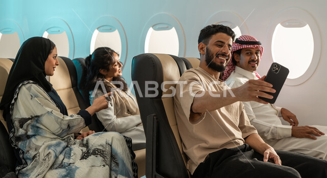 رحلات سياحية شبابية ، مجموعة من المسافرون العرب الخليجيون السعوديون في الطائرة ، التقاط صور تذكارية بإستخدام الهاتف المحمول ، رحلات ممتعة مع الاصدقاء ، الخطوط الجوية السعودية.
