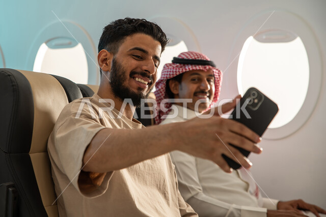 صديقان عربيان سعوديان  خليجيان يجلسان في مقعد الطائرة ، التقاط صور تذكارية بإستخدام الهاتف المحمول ، ايماءات تدل على السعادة والفرح ، اجواء ممتعة على متن الطائرة ، اجازة صيفية سعيدة ، الخطوط الجوية السعودية.