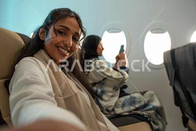 سيدتان عربيتان سعوديتان خليجيتان  تجلسان في مقعد الطائرة ، التقاط صور تذكارية بإستخدام الهاتف المحمول ، عطلة صيفية سعيدة  ، الخطوط الجوية السعودية ، ايماءات تدل على السعادة والفرح ، اجواء ممتعة على متن الطائرة.