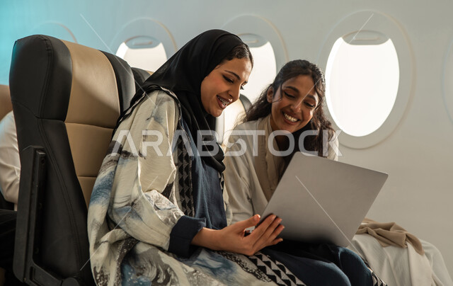 A tourist trip with friends, two Saudi Arabian Gulf women in the plane seat, summer vacation, using a laptop in the plane cabin, Saudi Airlines, tourism and travel, spending fun times with friends.