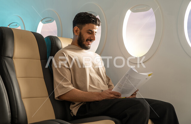  شاب عربي خليجي سعودي يرتدي اللباس الكاجوال يجلس في مقعد الطائرة ، ممارسة هواية القراءة ، قضاء أوقات ممتعة في مقصورة الطائرة ، قراءة الكتب المفيدة للتعلم والتثقف ، رحلة عمل ، خطوط الطيران السعودي.