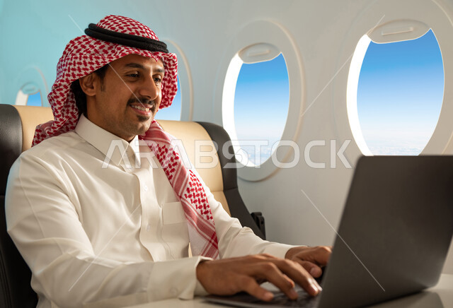 استخدام الحاسوب المحمول ، رجل عربي خليجي سعودي يرتدي الزي السعودي التقليدي يجلس في مقعد الطائرة ، انجاز مهام العمل في مقصورة الطائرة ، ايماءات تدل على السعادة والراحة ، الخطوط الجوية السعودية.