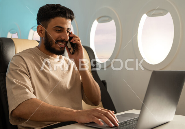  انجاز مهام العمل في مقصورة الطائرة  ، شاب عربي خليجي سعودي يرتدي اللباس الكاجوال ، يجلس في مقعد الطائرة ، اجراء مكالمة هاتفية ، استخدام الحاسوب المحمول ، الخطوط الجوية السعودية ، درجة رجال الاعمال.