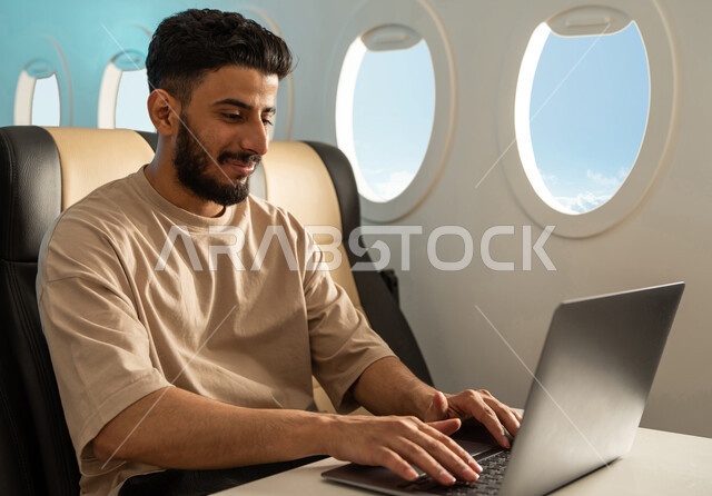 شاب عربي خليجي يرتدي اللباس الكاجوال ، تصفح الانترنت ، استخدام جهاز الحاسوب المحمول ، الخطوط الجوية السعودية ، رحلات جوية سياحية ،  انجاز مهام العمل في مقصورة الطائرة ، سياحة وسفر ، رحلة عمل.