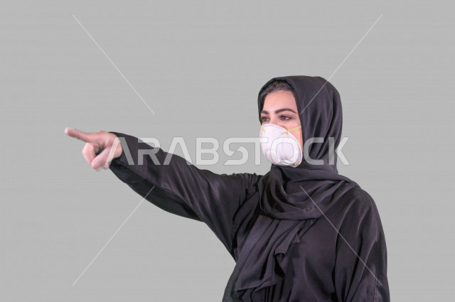 بورتريه لمرأة عربية خليجية سعودية ترتدي الكمامة والقفازات، اتخاذ اجراءات الوقاية من فايروس كورونا (كوفيد-19)، امرأة تشير بإصبعها السبابة لشيء ما، تجنب انتشار العدوى، خلفية بيضاء