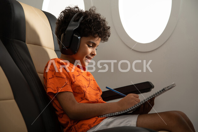 ارتداء سماعة رأس لا سلكية، طفل مسافر عربي خليجي سعودي يرتدي ملابس كاجوال ويقضي الوقت في الرسم والتلوين، الجلوس في مقعد الطائرة والإستماع الى الموسيقى، ايماءات وجه تدل على السعادة، رحلة سياحية من خلال خطوط الطيران السعودي