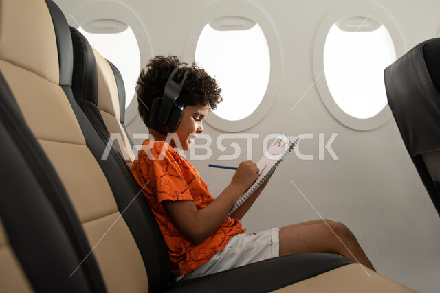 قضاء الوقت في الرسم والتلوين، ارتداء ملابس كاجوال والجلوس في مقعد الطائرة، طفل مسافر عربي خليجي سعودي يرتدي سماعة رأس لا سلكية ويستمع الى الموسيقى، ايماءات وجه تدل على السعادة، رحلة سياحية من خلال خطوط الطيران السعودي