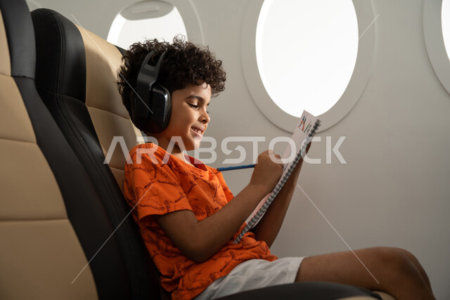 ارتداء سماعة رأس لا سلكية والإستماع الى الموسيقى، طفل مسافر عربي خليجي سعودي يرتدي ملابس كاجوال ويجلس في مقعد الطائرة، قضاء الوقت في الرسم والتلوين، ايماءات وجه تدل على السعادة، رحلة سياحية من خلال خطوط الطيران السعودي