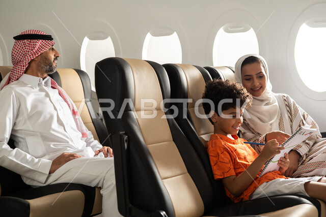 الاستعداد لقضاء العطلة الصيفية برفقة العائلة، عائلة عربية خليجية سعودية داخل الطائرة، طفل يقضي الوقت في الرسم والتلوين، رحلة سياحية عائلية من خلال خطوط الطيران السعودي