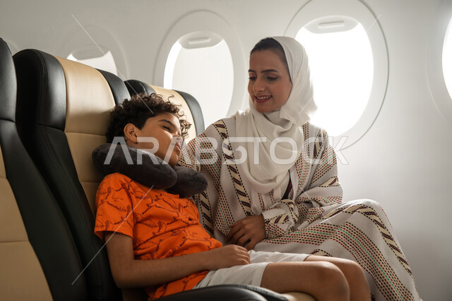 الشعور بالتعب والارهاق والحاجة الى النوم والراحة، امرأة عربية سعودية خليجية برفقة ولدها النائم في الطائرة، قضاء العطلة الصيفية برفقة العائلة، رحلة سياحية عائلية، خطوط الطيران السعودي