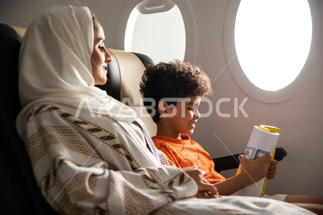 قضاء العطلة الصيفية برفقة العائلة، طفل يقضي وقت الرحلة في قراءة الكتب والقصص، امرأة عربية سعودية خليجية برفقة ولدها في الطائرة، رحلة سياحية عائلية، خطوط الطيران السعودي