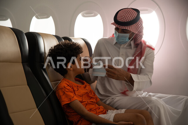 ولد عربي خليجي سعودي برفقة والده في الطيارة، ارتداء الكمامة الواقية والالتزام باجراءات الوقاية من فايروس كورونا، قضاء العطلة الصيفية برفقة العائلة، رحلة سياحية عائلية، خطوط الطيران السعودي