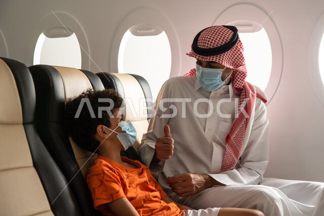 ارتداء الكمامة الواقية والالتزام باجراءات الوقاية من فايروس كورونا، ولد عربي خليجي سعودي برفقة والده في الطيارة، رجل يشير بإصبعه الإبهام لأعلى، القبول والموافقة والإعجاب، قضاء العطلة الصيفية برفقة العائلة، رحلة سياحية عائلية، خطوط الطيران السعودي