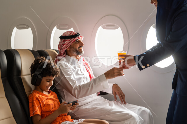 التسلة والترفيه باستخدام الهاتف المحمول، مضيفة الطيران تقوم بتقديم العصير لرجل عربي خليجي سعودي برفقة ولده في رحلة سياحية في الطائر، حسن الضيافة والاستقبال، طفل يرتدي سماعة رأس لا سلكية، ايماءات وجه تدل على السعادة، خطوط الطيران السعودي