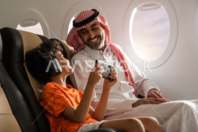 A child wearing a wireless headphone and listening to music, a Saudi Arabian Gulf man accompanied by his son on a tourist trip in Al-Tair, entertainment and entertainment using a mobile phone, facial gestures indicating happiness, spending the summer vaca