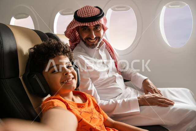 طفل يرتدي سماعة رأس لا سلكية ويستمع الى الموسيقى، رجل عربي خليجي سعودي برفقة ولده في رحلة سياحية في الطائرة، ايماءات وجه تدل على السعادة، قضاء العطلة الصيفية برفقة العائلة، خطوط الطيران السعودي