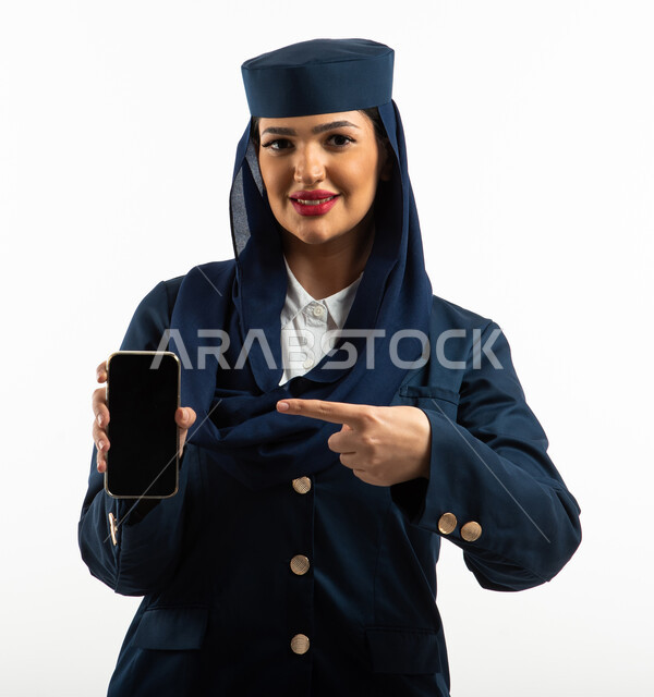بورتريه لمضيفة طيران عربية خليجية سعودية مبتسمة ترتدي زي مضيفات الطيران ، الاشارة الى شاشة سوداء مفرغه على الهاتف المحمول ، استخدام تطبيقات وبرامج الجوال في مجال الطيران , خطوط الطيران السعودي ، خلفية بيضاء ، مهنة مضيفة الطيران ، طاقم الطائرة.