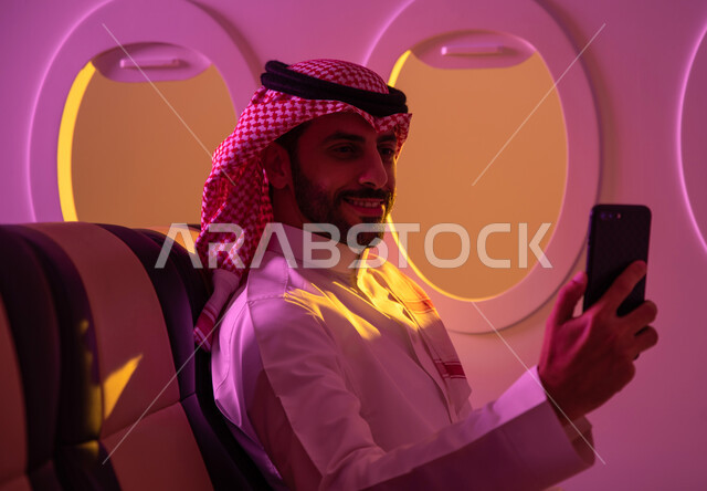 Means of air transportation, wearing traditional Saudi dress, a Saudi Arabian Gulf man sitting in the cabin of the plane, taking selfies via mobile phone, tourist flights, Saudi Airlines.