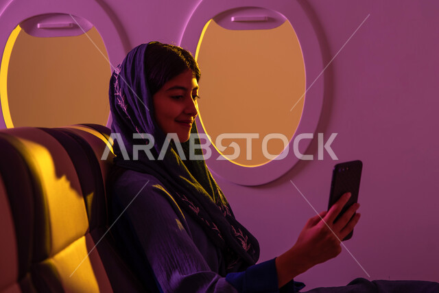 قضاء عطلة صيفية سياحية, ايماءات تدل على السعادة, التقاط صور سيلفي عبر الهاتف المحمول, امرأة عربية  سعودية خليجية تجلس في الطائرة , ايماءات تدل على السعادة, رحلات جوية سياحية, الخطوط الجوية السعودية, وسائل مواصلات جوية .