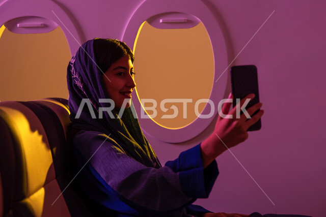 قضاء عطلة صيفية ممتعة, ايماءات تدل على السعادة, التقاط صور سيلفي عبر الهاتف المحمول, امرأة عربية  سعودية خليجية تجلس في الطائرة , ايماءات تدل على السعادة, رحلات جوية سياحية, الخطوط الجوية السعودية, وسائل مواصلات جوية .