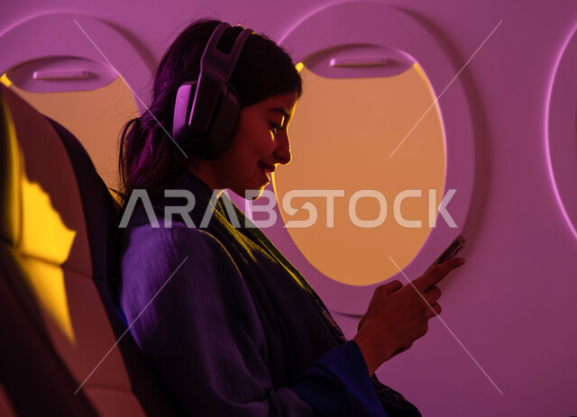 وسائل نقل جوية, خطوط جوية سعودية, عطلة صيفية سياحية, ايماءات تدل على السعادة, التواصل الاجتماعي, امرأة عربية خليجية سعودية تجلس في الطائرة , رحلات جوية سياحية, استخدام تقنية الهيدفون, استخدام الهاتف المحمول .