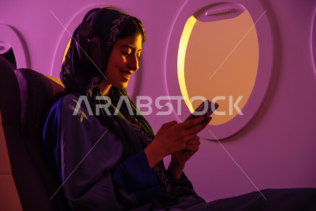 خطوط جوية سعودية, عطلة صيفية سياحية, ايماءات تدل على السعادة, التواصل الاجتماعي, امرأة عربية خليجية سعودية تجلس في الطائرة , رحلات جوية سياحية, استخدام تقنية الهيدفون, استخدام الهاتف المحمول, وسائل مواصلات جوية .