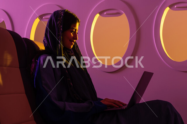  انجاز مهام العمل عبر جهاز الحاسوب المحمول ، ايماءات تدل على السعادة, رحلة عمل سعيدة ،امرأة سعودية عربية خليجية  محجبة ترتدي العباءة الخليجية ، الجلوس في مقصورة الطائرة ، الخطوط الجوية السعودية.