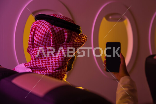 رحلات جوية سياحية, ارتداء الزي السعودي التقليدي, عرض شاشة سوداء فارغة عبر الهاتف المحمول, صورة من الخلف لرجل عربي خليجي سعودي يجلس في مقعد الطائرة, تصفح الانترنت ، خطوط الطيران السعودي ،رحلة عمل.