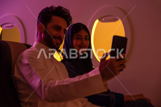 التقاط صور السيلفي عبر الهاتف المحمول , ايماءات تدل على الفرح والسرور, زوجان عربيان سعوديان خليجيان في مقعد الطائرة, دردشة ومحادثات, عطلة صيفية سعيدة, درجة رجال أعمال, الخطوط الجوية السعودية, رحلة عمل, سياحة وسفر, رحلات جوية سياحية .