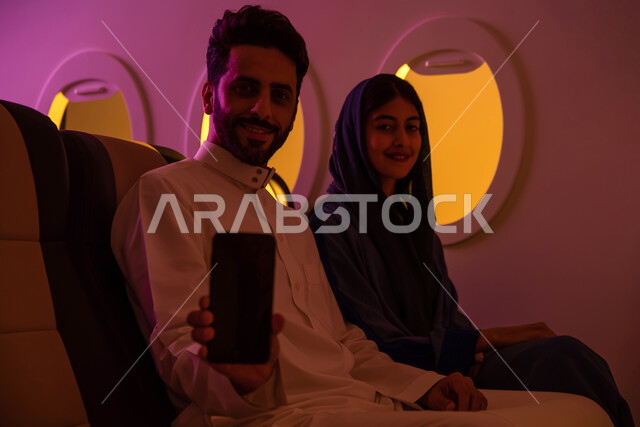عرض شاشة سوداء فارغة على الهاتف المحمول, ايماءات تدل السرور والفرح, عطلة صيفية سعيدة, زوجان عربيان سعوديان خليجيان في مقعد الطائرة, دردشة ومحادثات, الخطوط الجوية السعودية, رحلة عمل, سياحة وسفر, رحلات جوية سياحية .