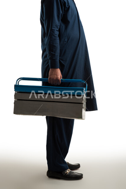 صورة مقربة من الجانب لعامل بناء يحمل بيده صندوق ادوات معدني، بورتريه لرجل باكستاني يرتدي الزي الباكستاني التقليدي، صندوق عدة حديدي، استقدام العمالة من الخارج، صورة الجسم بالطول الكامل، خلفية بيضاء