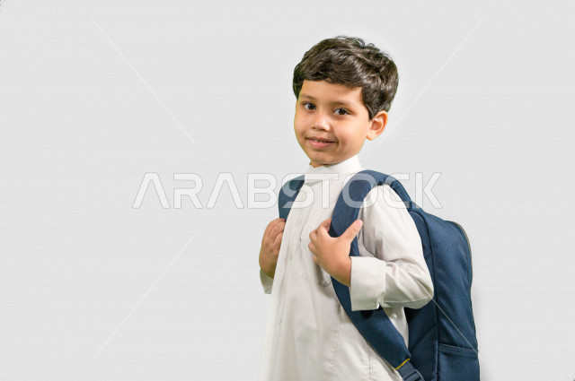 بورتريه لطالب مدرسي عربي خليجي سعودي يرتدي حقيبة ظهر مدرسية، ارتداء الزي المدرسي التقليدي، ايماءات وجه ويدين تدل على السعادة، خلفية بيضاء