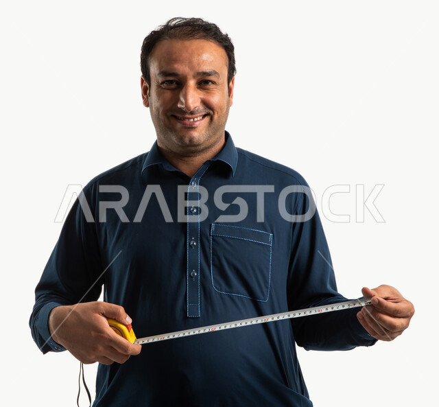 بورتريه لعامل بناء باكستاني بايماءات وجه سعيدة يرتدي الزي الباكستاني التقليدي ويمسك بيده اداة قياس الطول، مقياس شريطي، علامات القياس، ادوات التشطيب، مهن وحرف يدوية، استقدام العمالة من الخارج، اتقان العمل، خلفية بيضاء