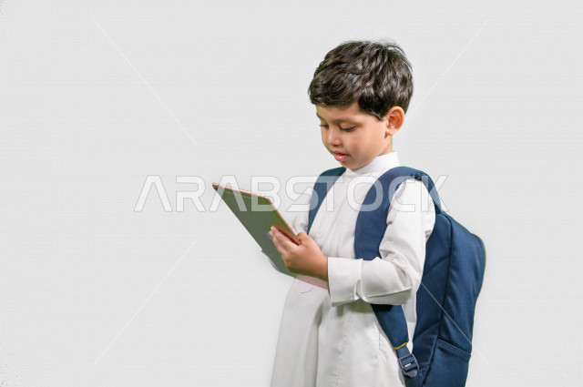 بورتريه لطالب مدرسي عربي خليجي سعودي يرتدي حقيبة ظهر مدرسية، ارتداء الزي المدرسي التقليدي، استخدام التابليت المحمول في التعلم، التعلم عن بعد، التعليم الالكتروني، خلفية بيضاء