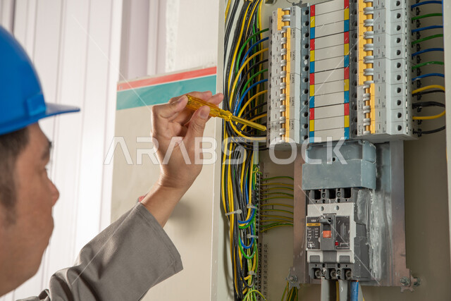 عامل صيانة كهربائي يرتدي خوذة الحماية ويستخدم مفك فحص الكهرباء، صيانة وتركيب لوحة عداد الكهرباء، مفهوم الهندسة الكهربائية، تركيب مفاتيح الكهرباء، عمل فني كهربائي، تمديد اسلاك الكهرباء، استقدام العمالة من الخارج