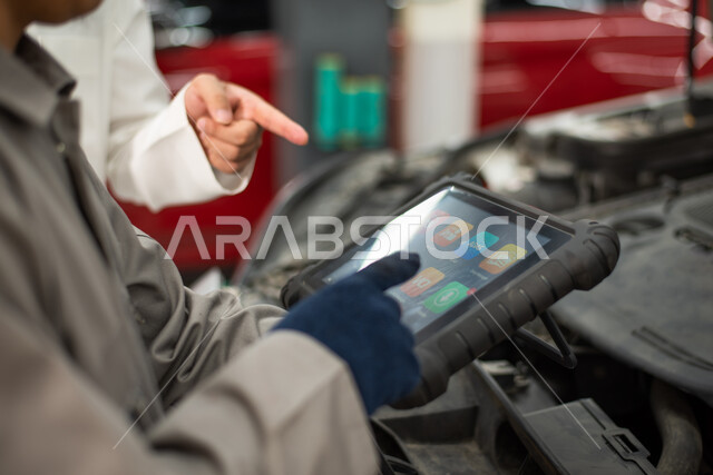 صورة مقربة لعامل صيانه ميكانيكي ورجل عربي سعودي خليجي يتحاوران حول تصليح أعطال اجزاء السياره، أعمال الصيانه و الفحص والتشييك على أجزاء السياره,  استخدام جهاز تقني متطور لاكتشاف العطل, حوار ونقاش بين عامل صيانه ميكانيكي ورجل عربي سعودي خليجي حول تصليح أعطا
