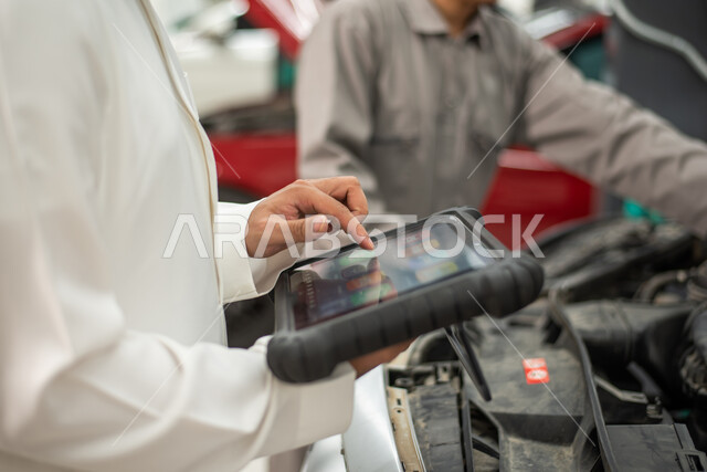 عامل صيانة يقوم باصلاح سيارة في الورشة, استخدام التقنية والتكنولوجيا لصيانة السياره في الورشه, طلب تصليح السياره عن طريق جهاز التابلت, صورة مقربة  لرجل عربي سعودي خليجي يرتدي الزي السعودي التقليدي يقف في مركز صيانة السيارات, مهندس ميكانيكي محترف