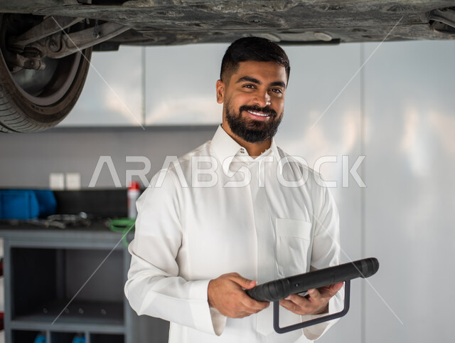 ايماءات تدل على السعادة, استخدام التقنية والتكنولوجيا لصيانة السياره في الورشه, طلب تصليح السياره عن طريق جهاز التابلت, رجل عربي سعودي خليجي يرتدي الزي السعودي التقليدي يقف في مركز صيانة السيارات, مهندس ميكانيكي محترف