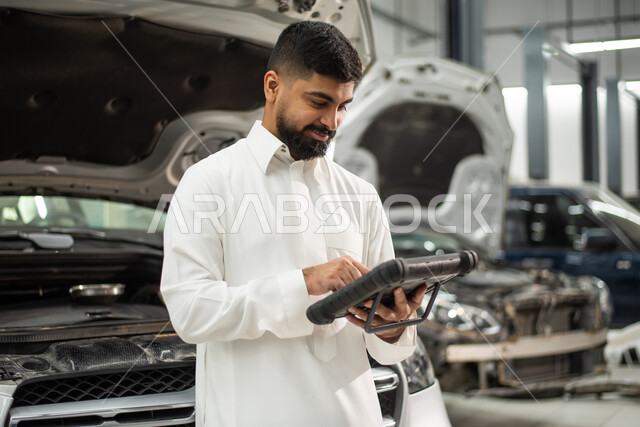 استخدام التقنية والتكنولوجيا لصيانة السياره في الورشه, طلب تصليح السياره عن طريق جهاز التابلت, رجل عربي سعودي خليجي يرتدي الزي السعودي التقليدي يقف في مركز صيانة السيارات, مهندس ميكانيكي محترف